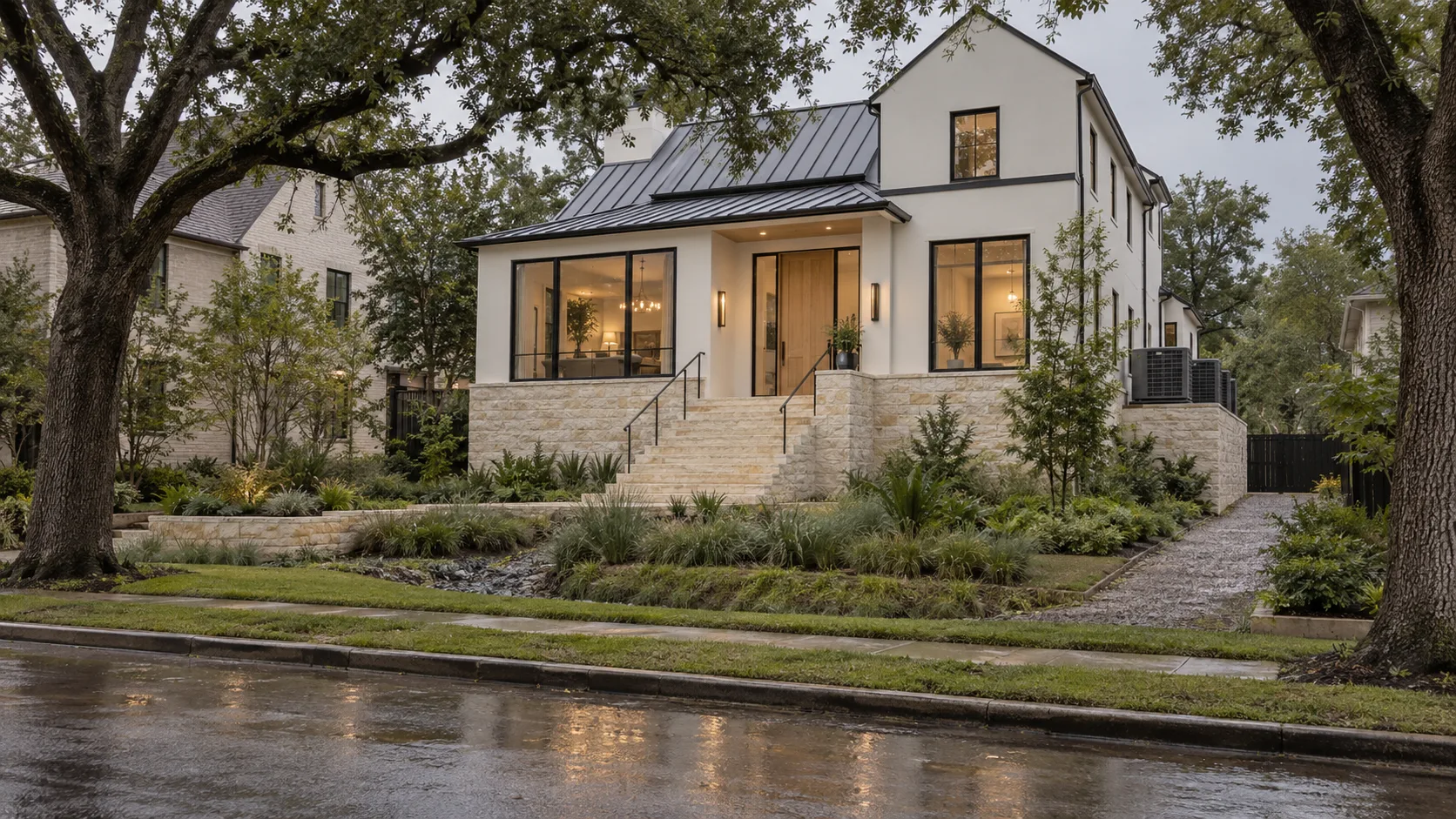 A two-story white plaster house with a dark standing seam metal roof and black-framed windows, its floor raised several feet above grade on a pale limestone base, with a wide flight of stone steps and two black steel handrails leading up to a white oak front door, air-conditioning condensers on a raised stone platform at the side, a rock-lined drainage swale through the planted front yard, live oaks overhead and a wet street in the foreground after rain