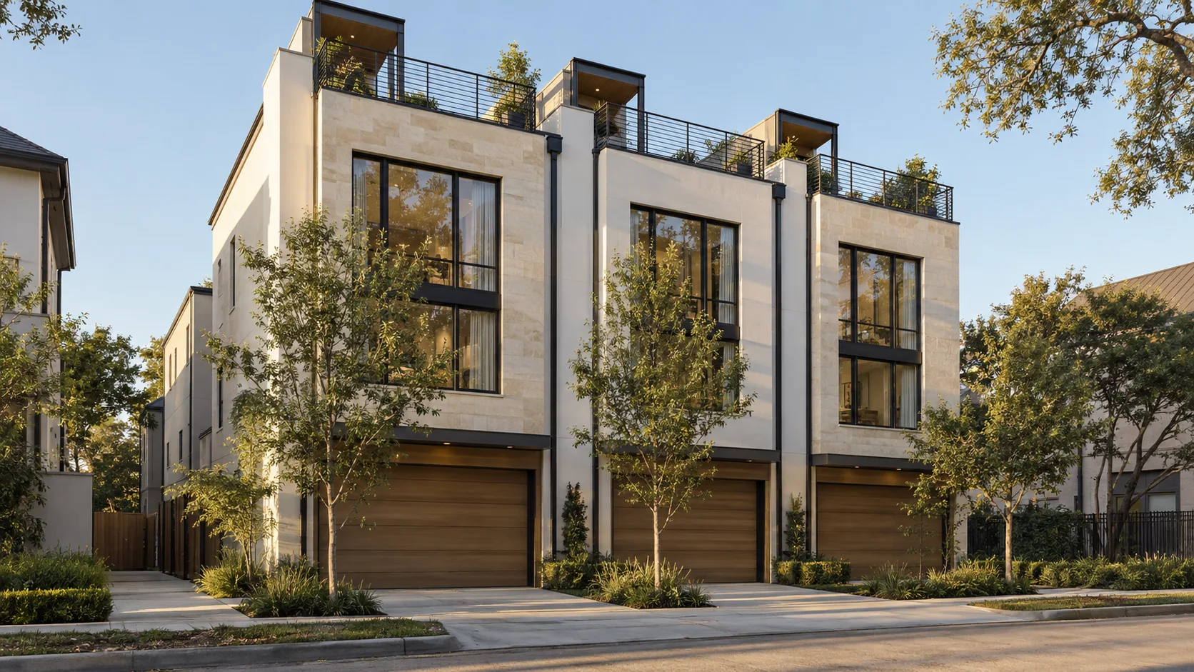 A row of 3 attached three-story townhouses faced in pale limestone and white plaster, each with a tall black-framed window stacked over a warm wood garage door, black steel downspouts between the units, rooftop terraces edged with black horizontal-bar steel railings and small covered stair enclosures with potted plants, young trees and planted beds along short concrete drives, in low late-afternoon sun with live oak branches at the top of the frame