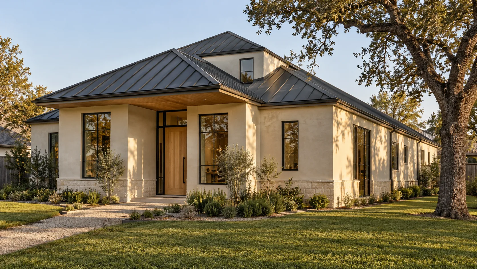 A warm modern Houston house with a charcoal standing-seam metal roof over a white oak soffit and limewashed plaster walls, with a hip roof, a live oak at the side and late-afternoon light